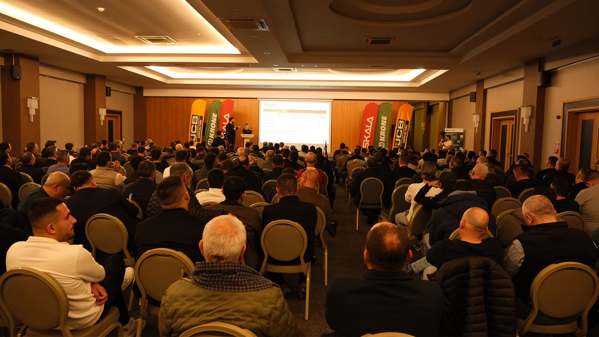 Large audience of men at a conference, facing a speaker and projected screen with JCB and Krone banners.