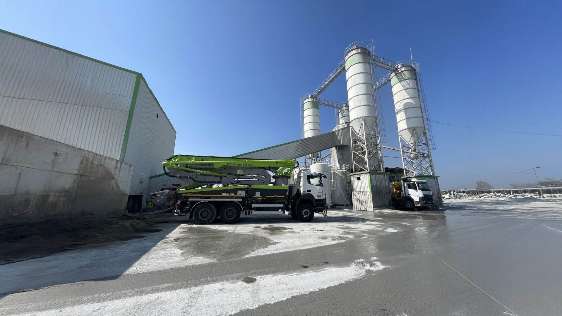 Concrete pump truck and batching plant at a large industrial facility under a clear sky.