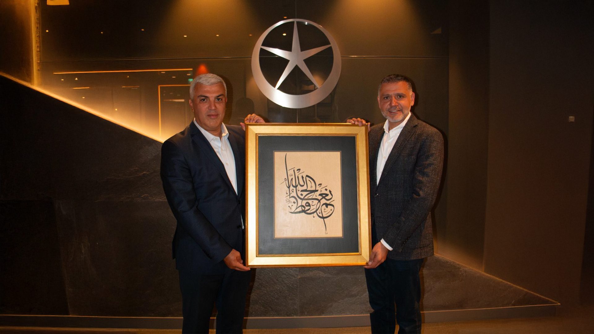 Two men holding a framed Arabic calligraphy artwork in front of a logo with a star.