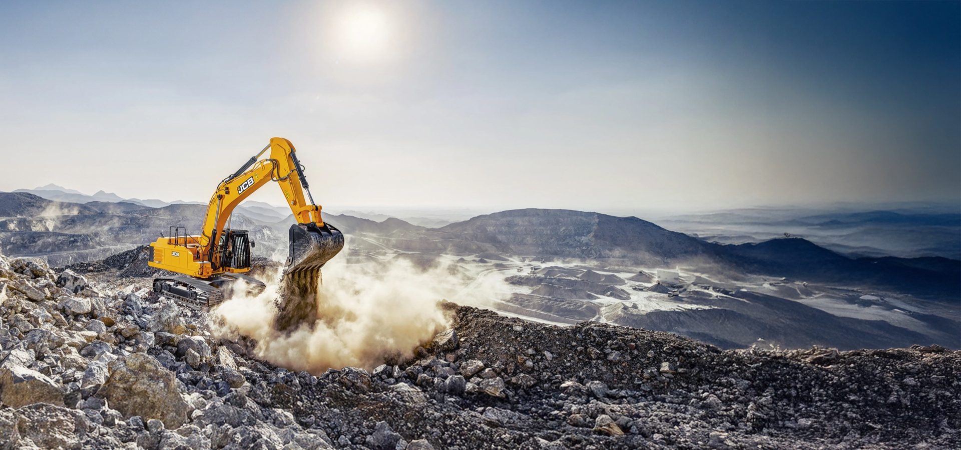 A yellow JCB excavator digs into a rocky terrain, kicking up dust, with a vast open-pit mine and mountains under a bright sun.