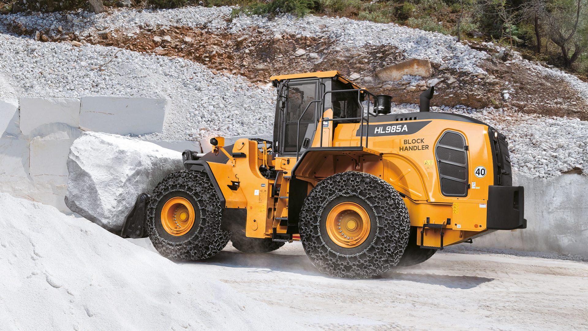 Yellow HL985A block handler with tire chains moving a large white rock in a quarry.
