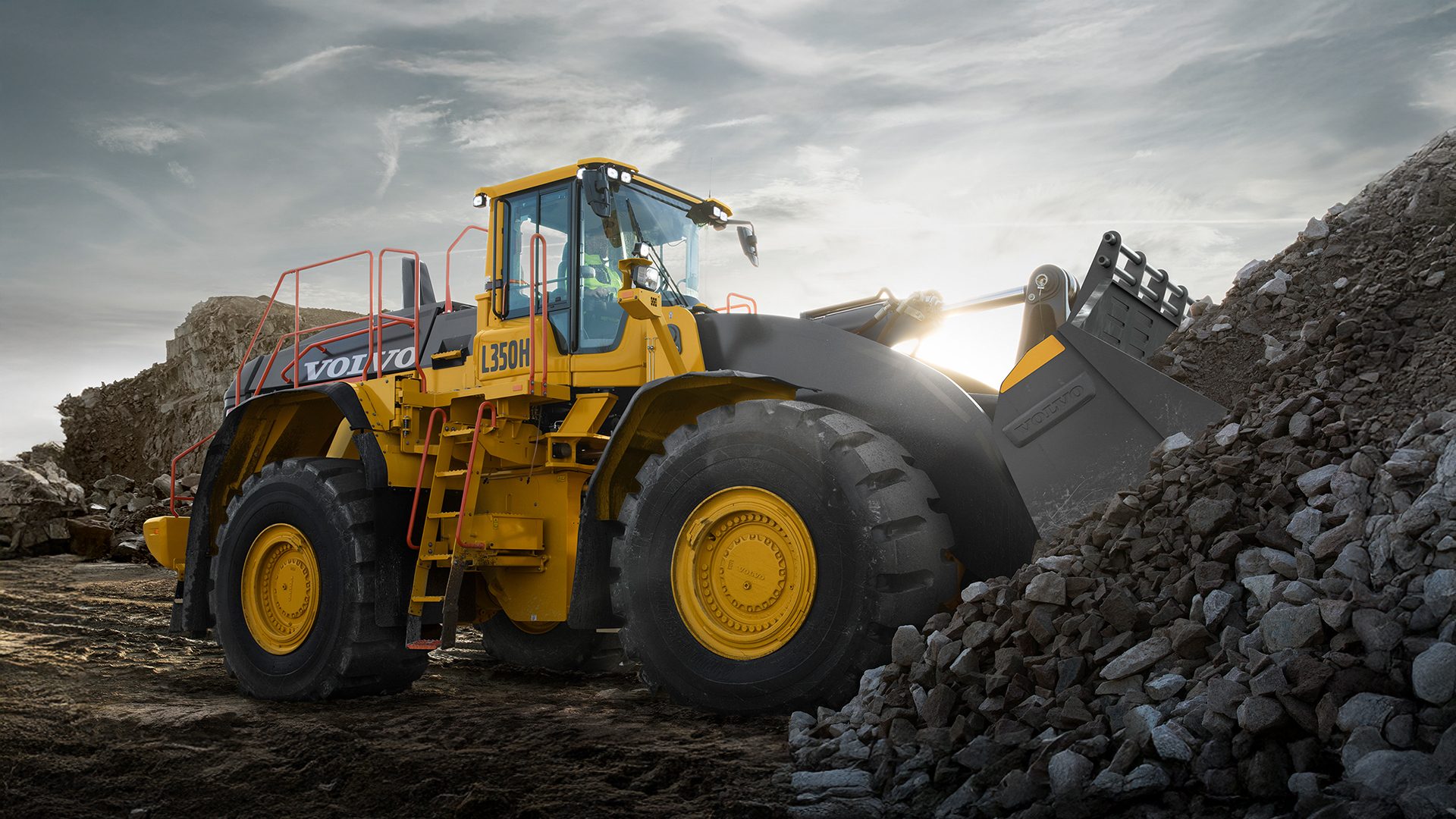 Yellow Volvo L350H wheel loader scooping rocks at a worksite under a cloudy sky.