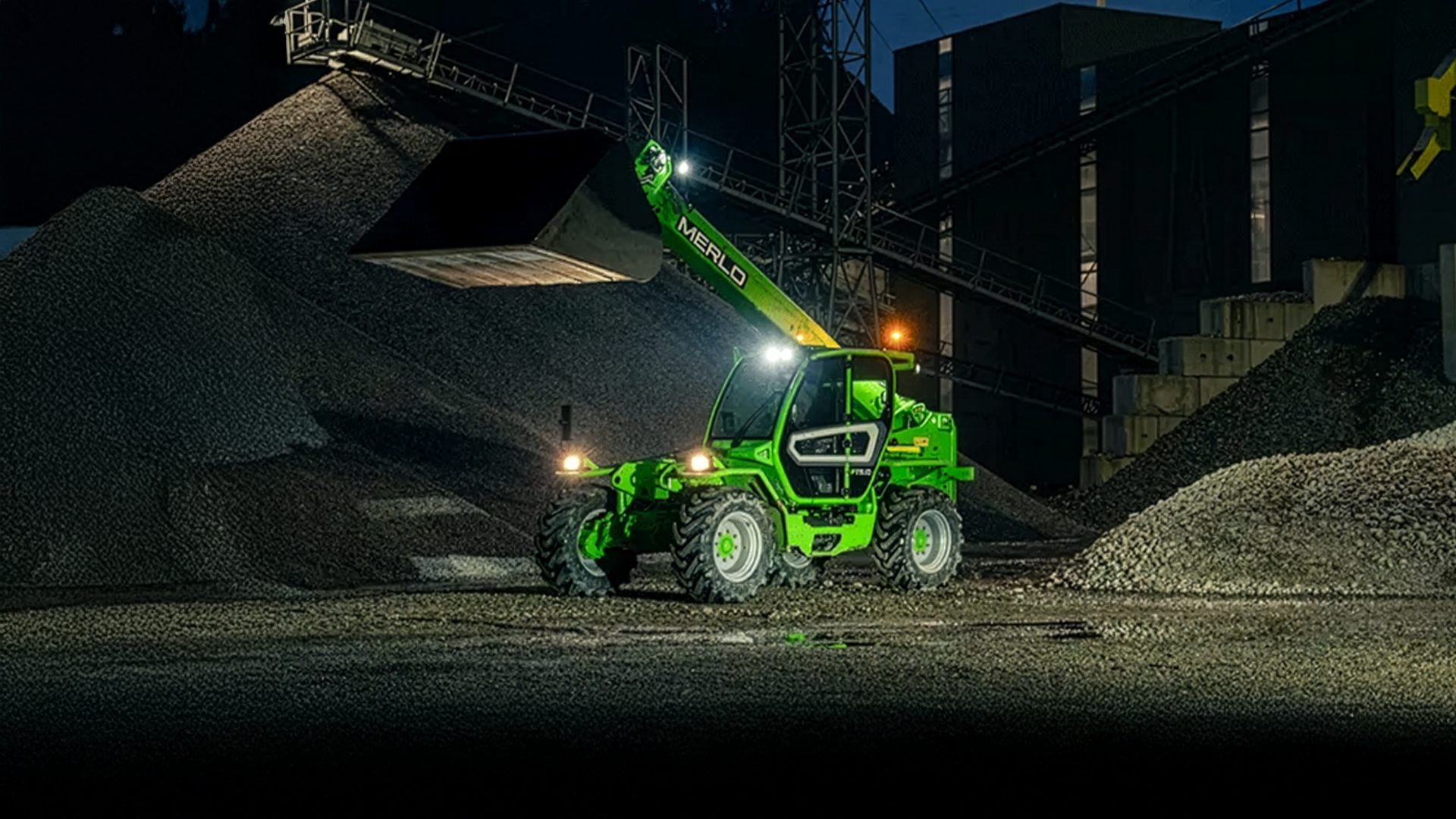 Green Merlo telehandler with lights on, loading gravel at a night construction site.