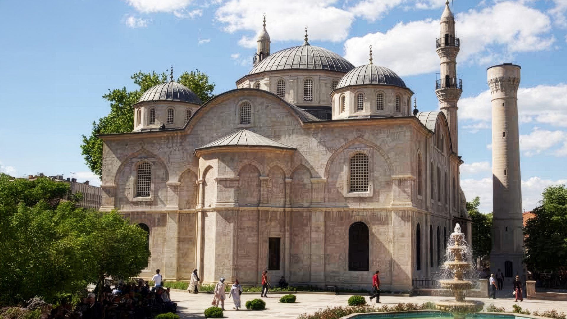 A large, light-colored mosque with multiple domes, two minarets, and a fountain on a sunny day.