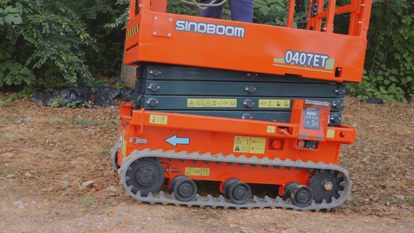 A vibrant orange Sinoboom 0407ET tracked scissor lift stands on a dirt path with lush green foliage.