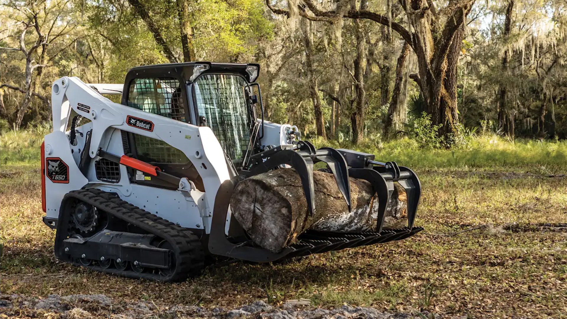 A Bobcat T650 compact track loader with a grapple attachment carries a large log in a wooded area.