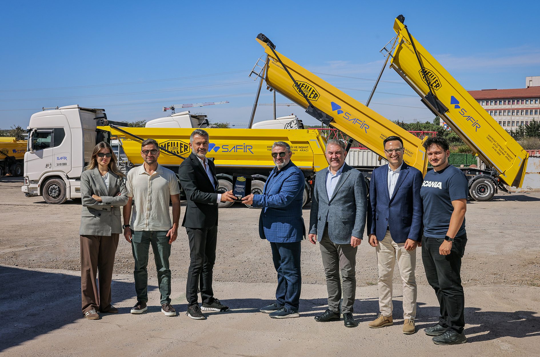 Seven people, two holding an award, pose with yellow dump trucks at an outdoor event.