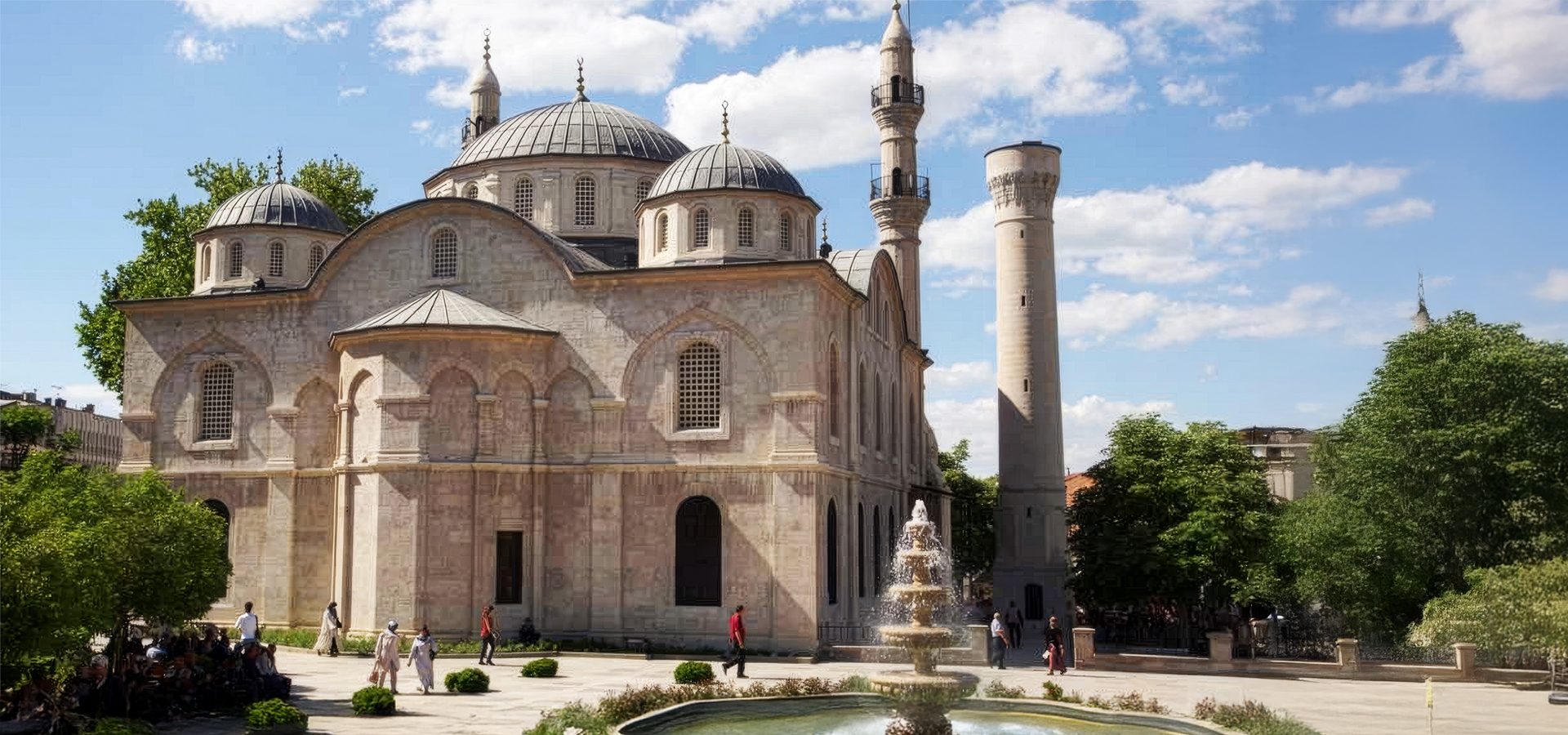 Grand mosque with domes, minaret, and a tiered fountain in a sunny courtyard with people.