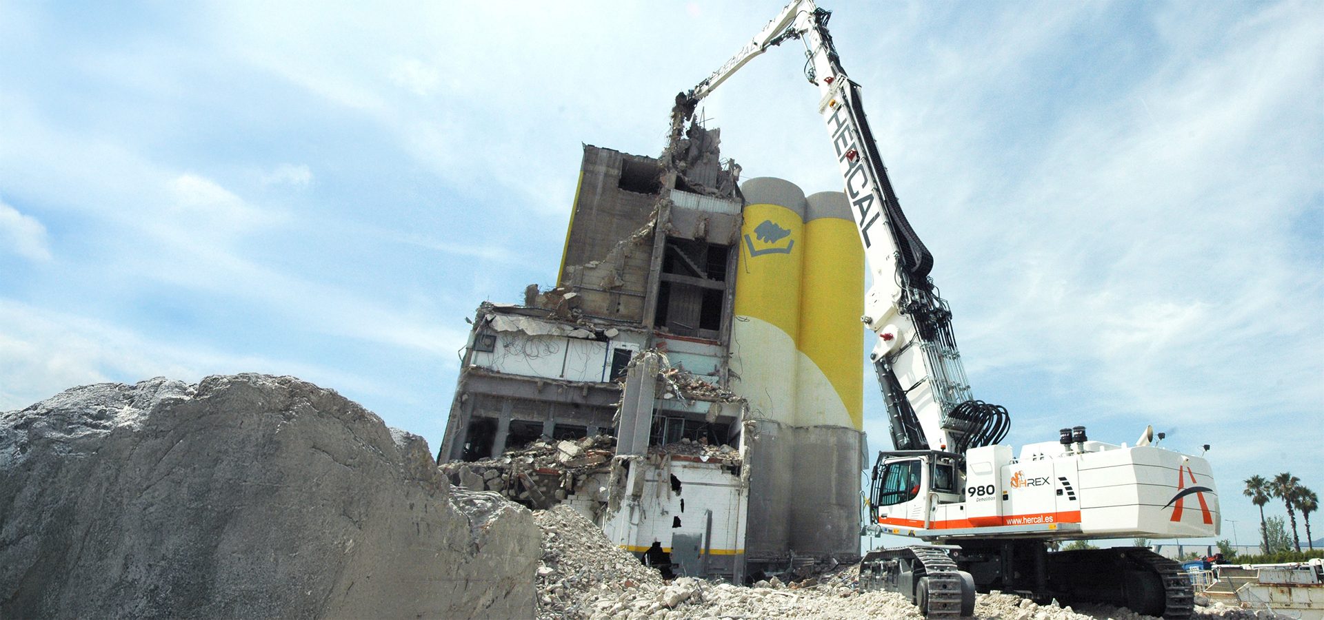 Large demolition excavator breaking down a concrete building next to yellow industrial silos.