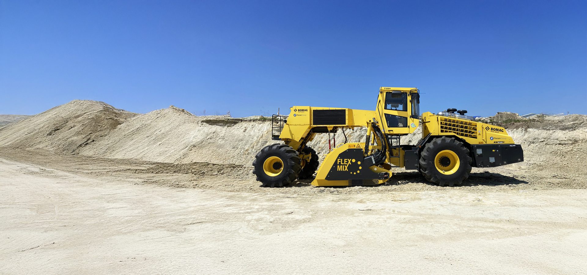 Large yellow BOMAG Flex Mix construction vehicle in a sandy quarry under a clear blue sky.