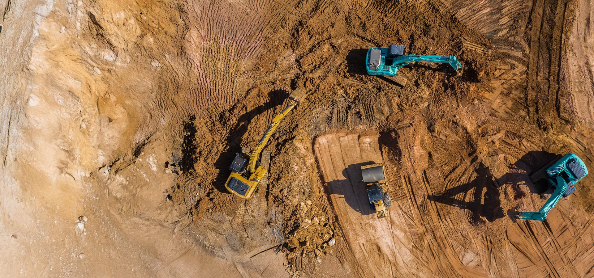 Aerial view of a construction site with multiple excavators and a roller working on brown soil.
