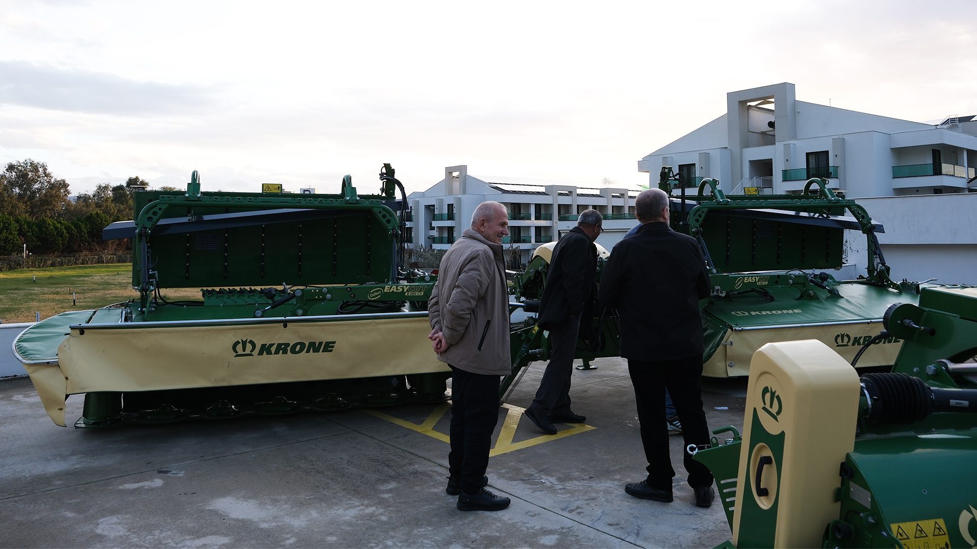 Men inspecting green Krone mowers outdoors, modern buildings in background.