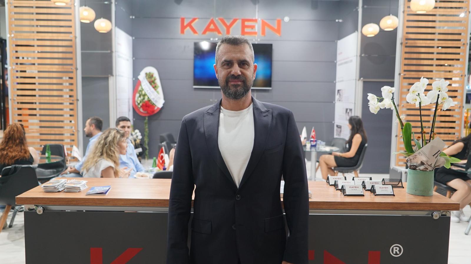 A man in a suit standing at a trade show booth with the "KAYEN" logo visible in the background.