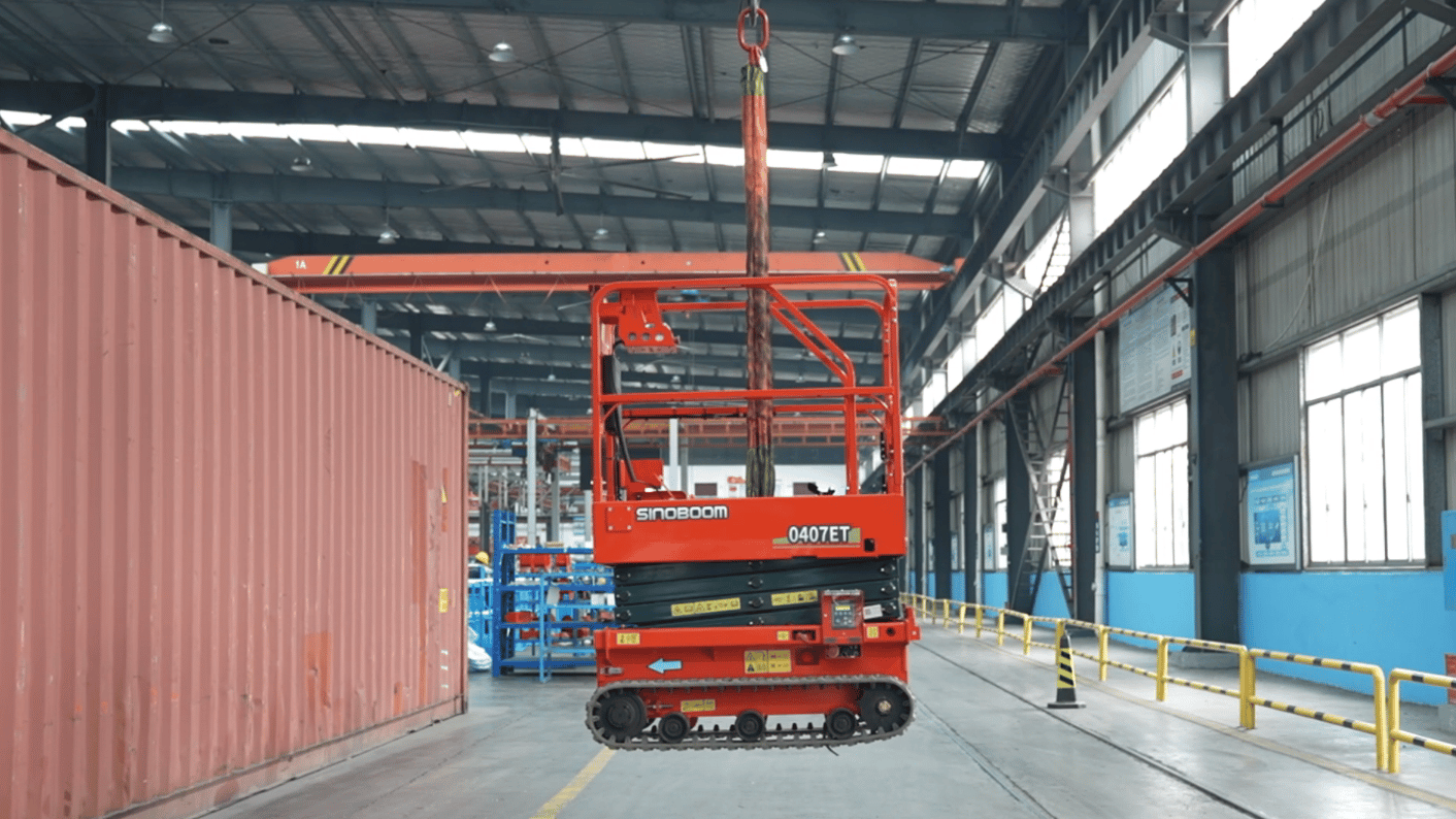 Red tracked scissor lift (SINOBOOM 0407ET) suspended in a large industrial building, with a cargo container to the left.