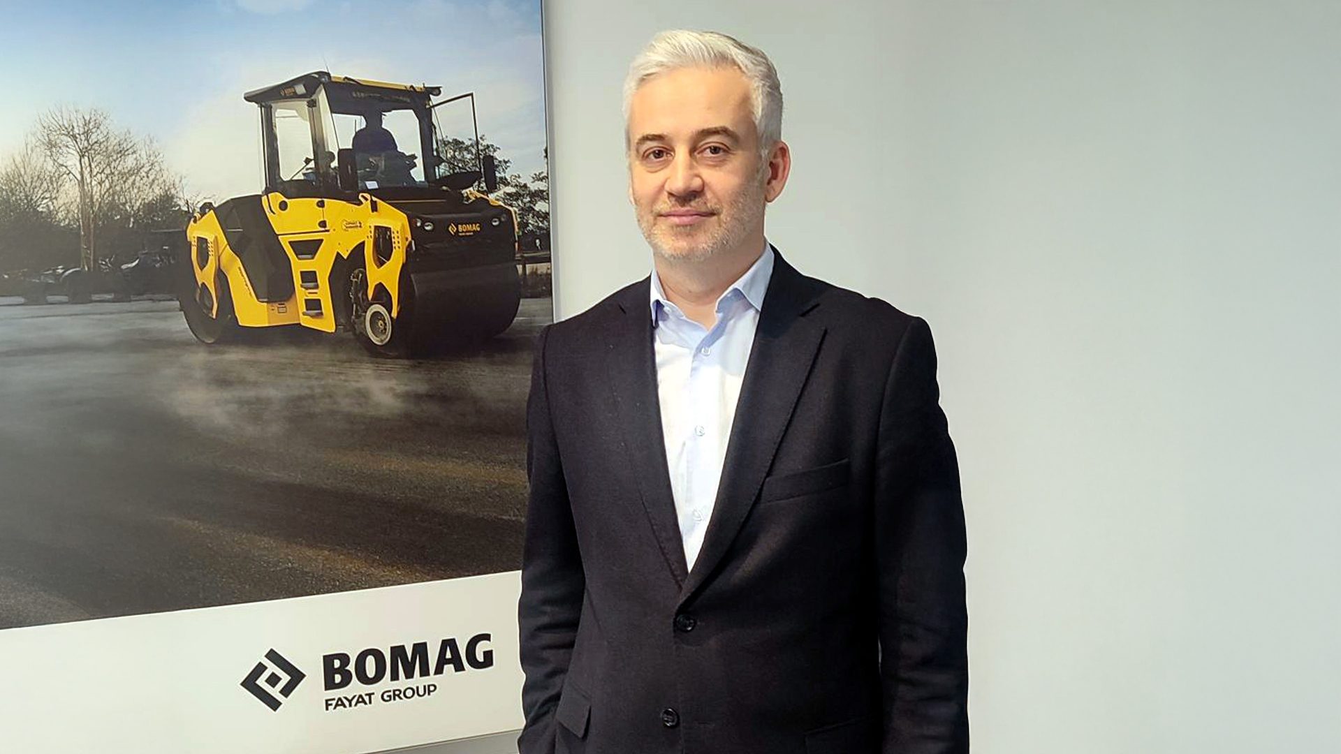 Man in a suit standing by a poster of a yellow BOMAG road roller.