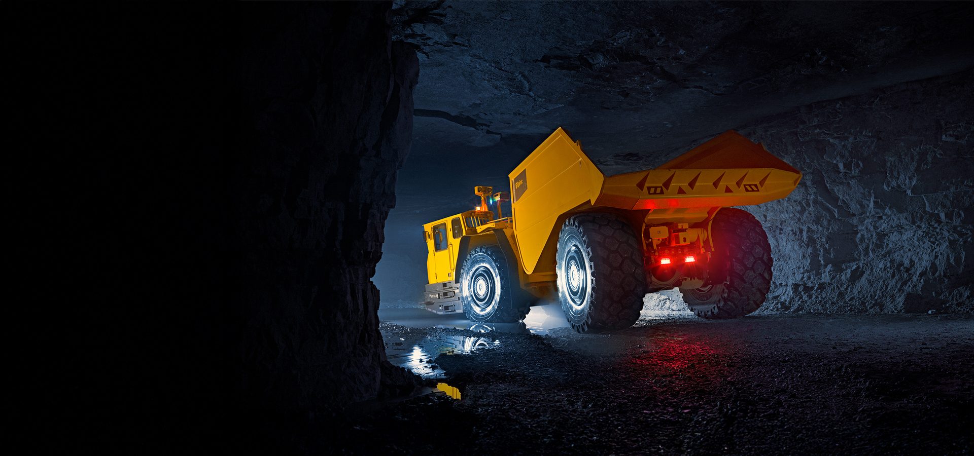 A large yellow mining dump truck with headlights and taillights on, inside a dark underground mine.