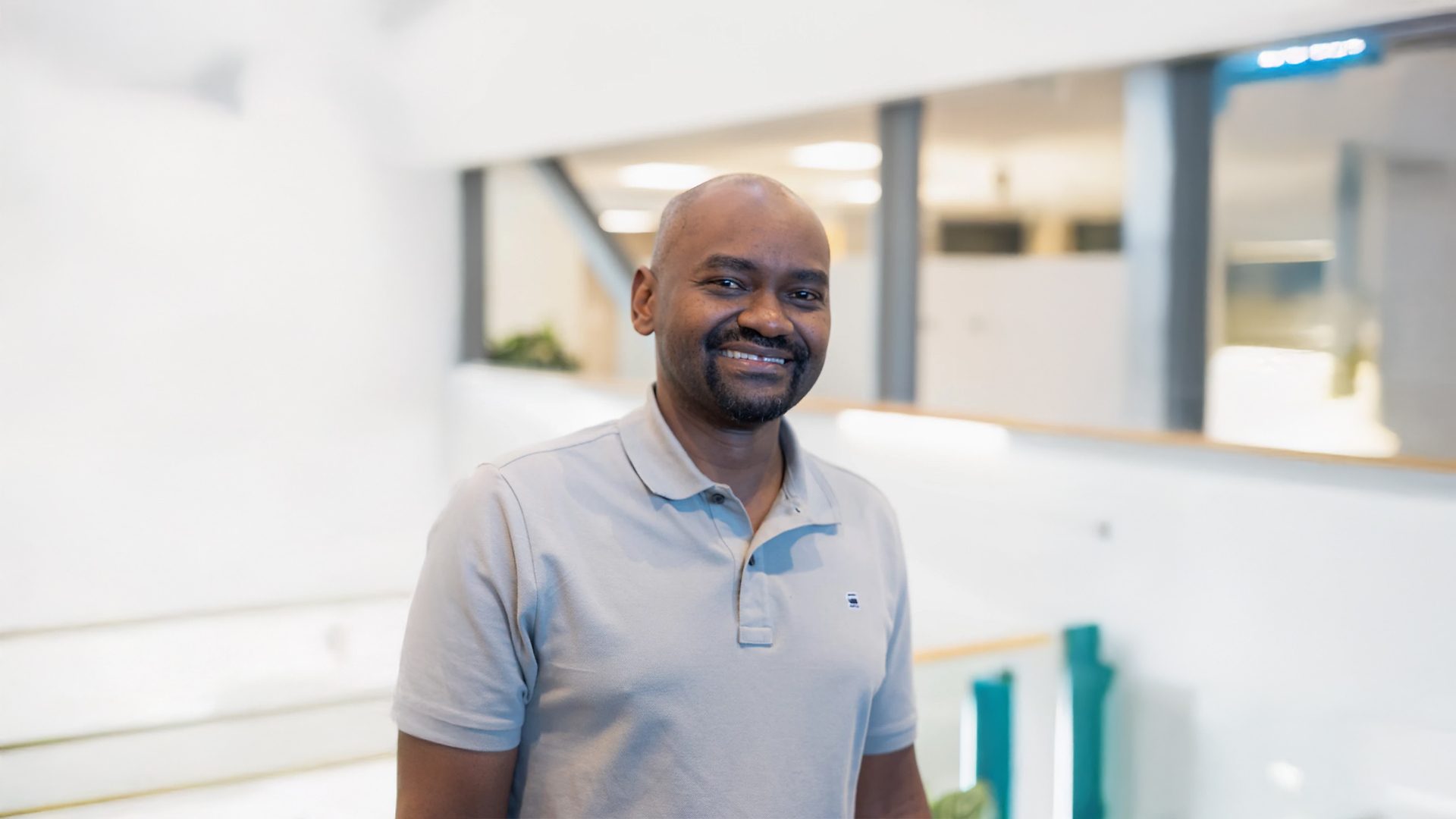 A smiling man with dark skin, wearing a grey polo shirt, stands indoors against a bright, modern background.