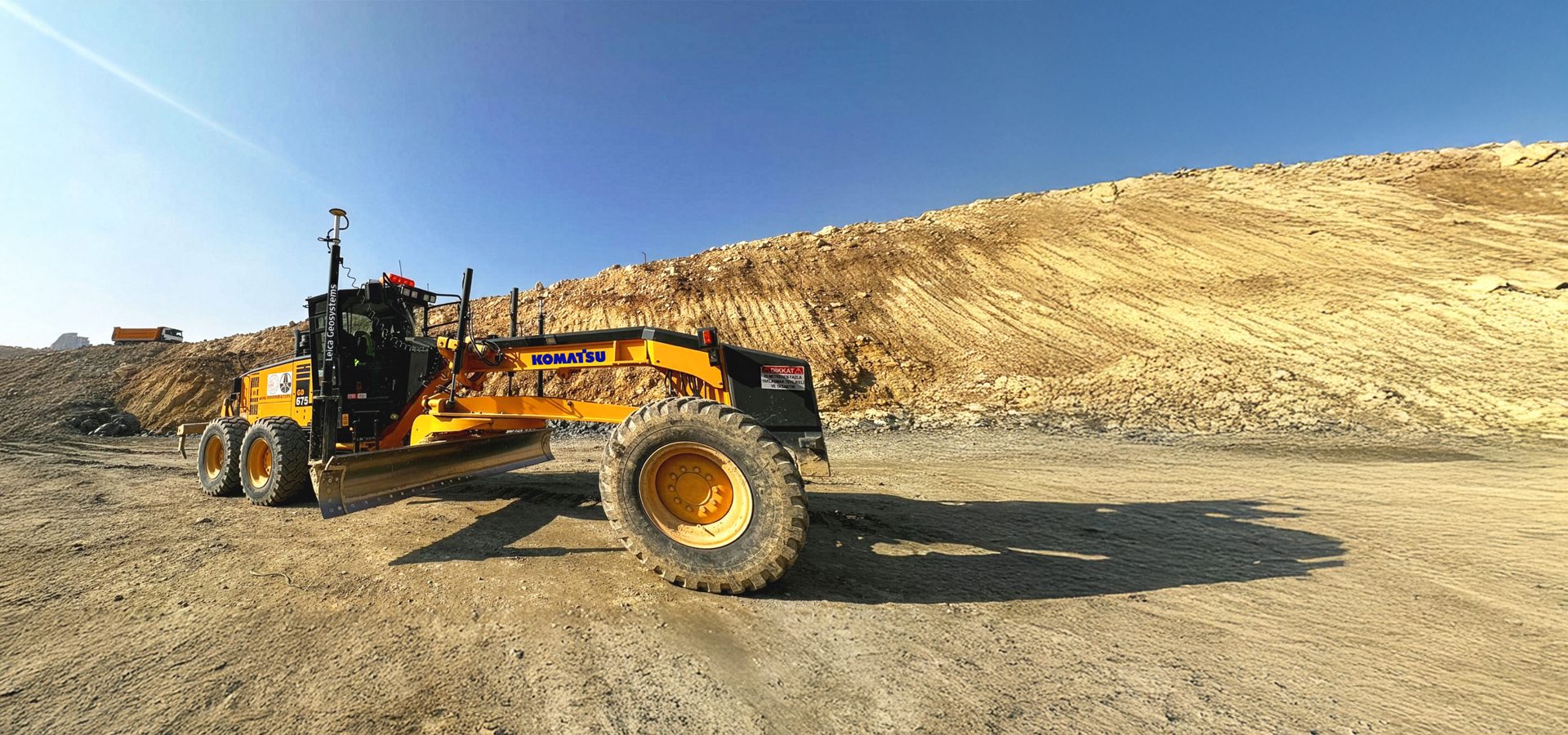 Yellow Komatsu grader at a dusty construction site with dirt piles under a clear blue sky.