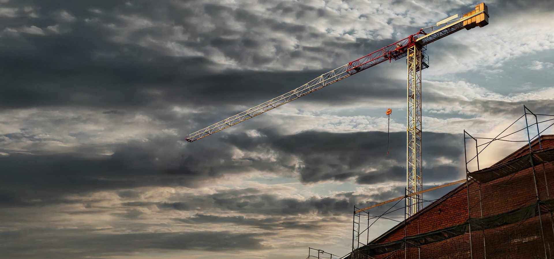 A large construction crane stands against a dramatic, dark cloudy sky, above a partially built brick building.