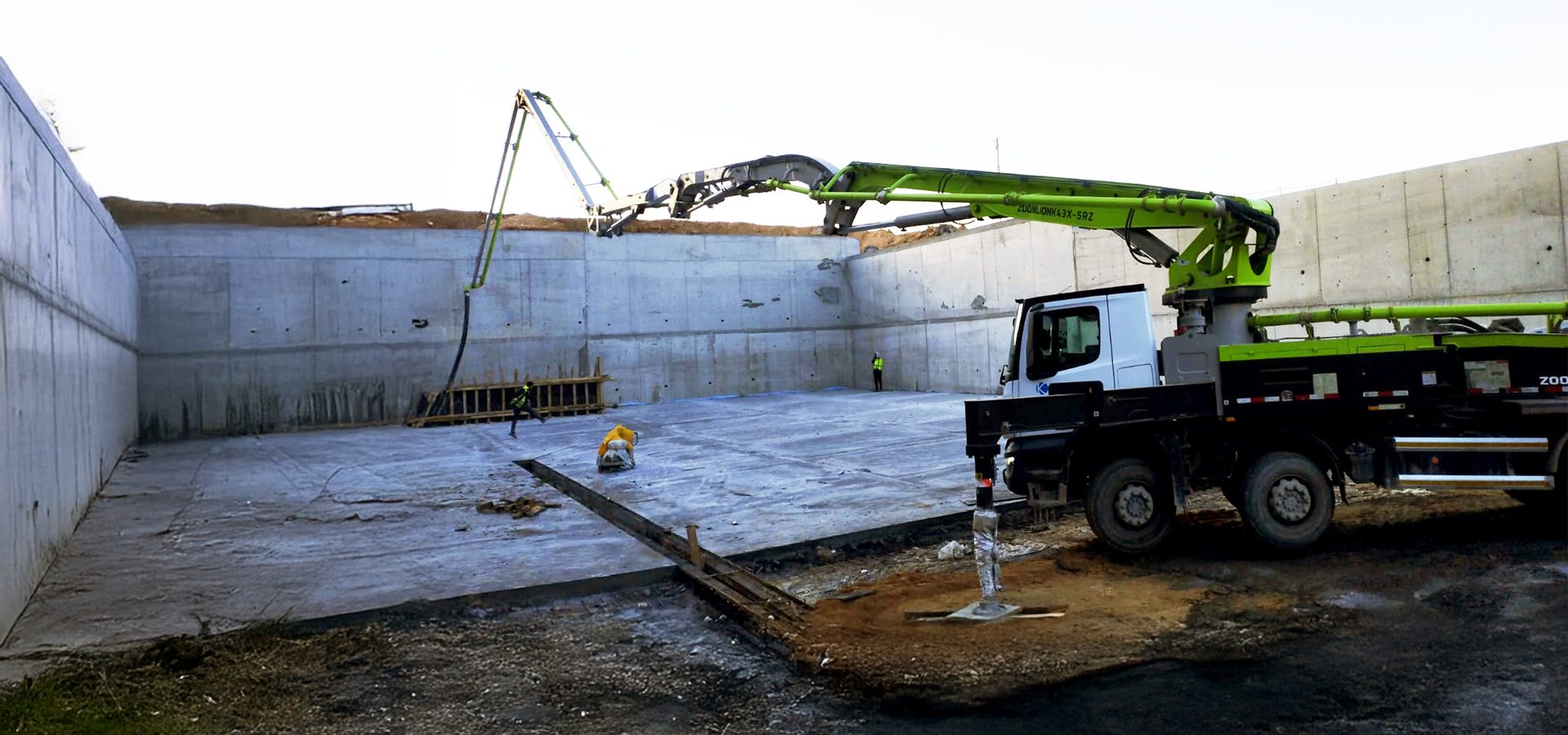 Concrete pump truck with extended boom pours concrete into a large walled foundation. Workers present.