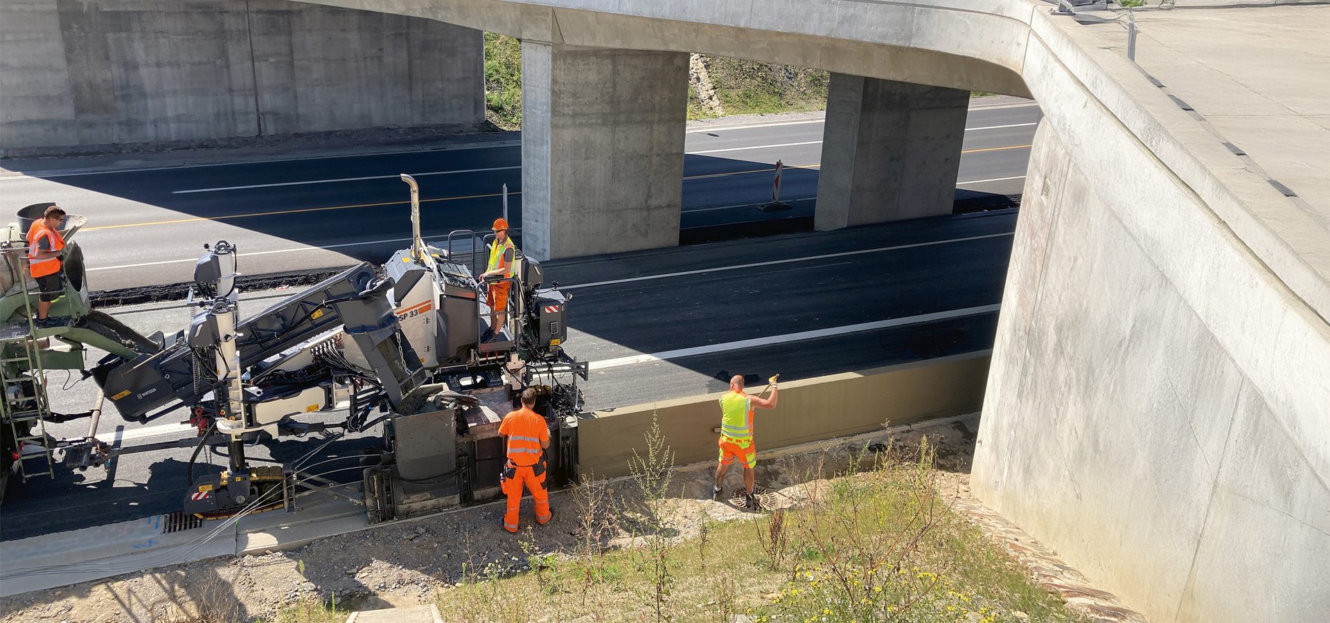 Workers operate a slipform paver laying concrete curb beside a highway under a bridge.