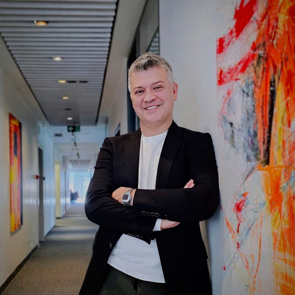 Smiling man in a black blazer and white t-shirt, leaning against a wall with abstract art in an office.