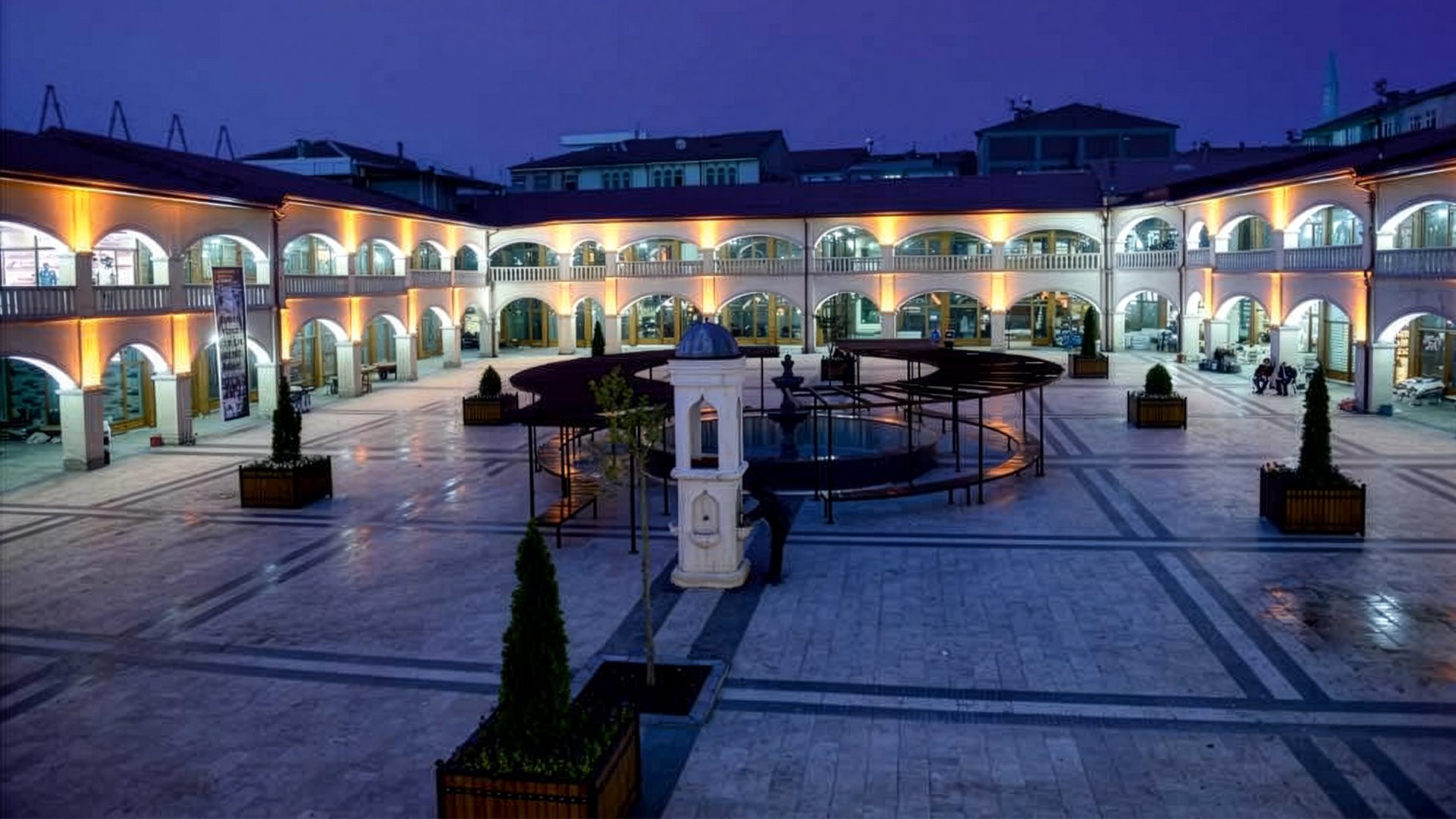Arched, illuminated buildings enclose a courtyard with a central fountain at twilight.