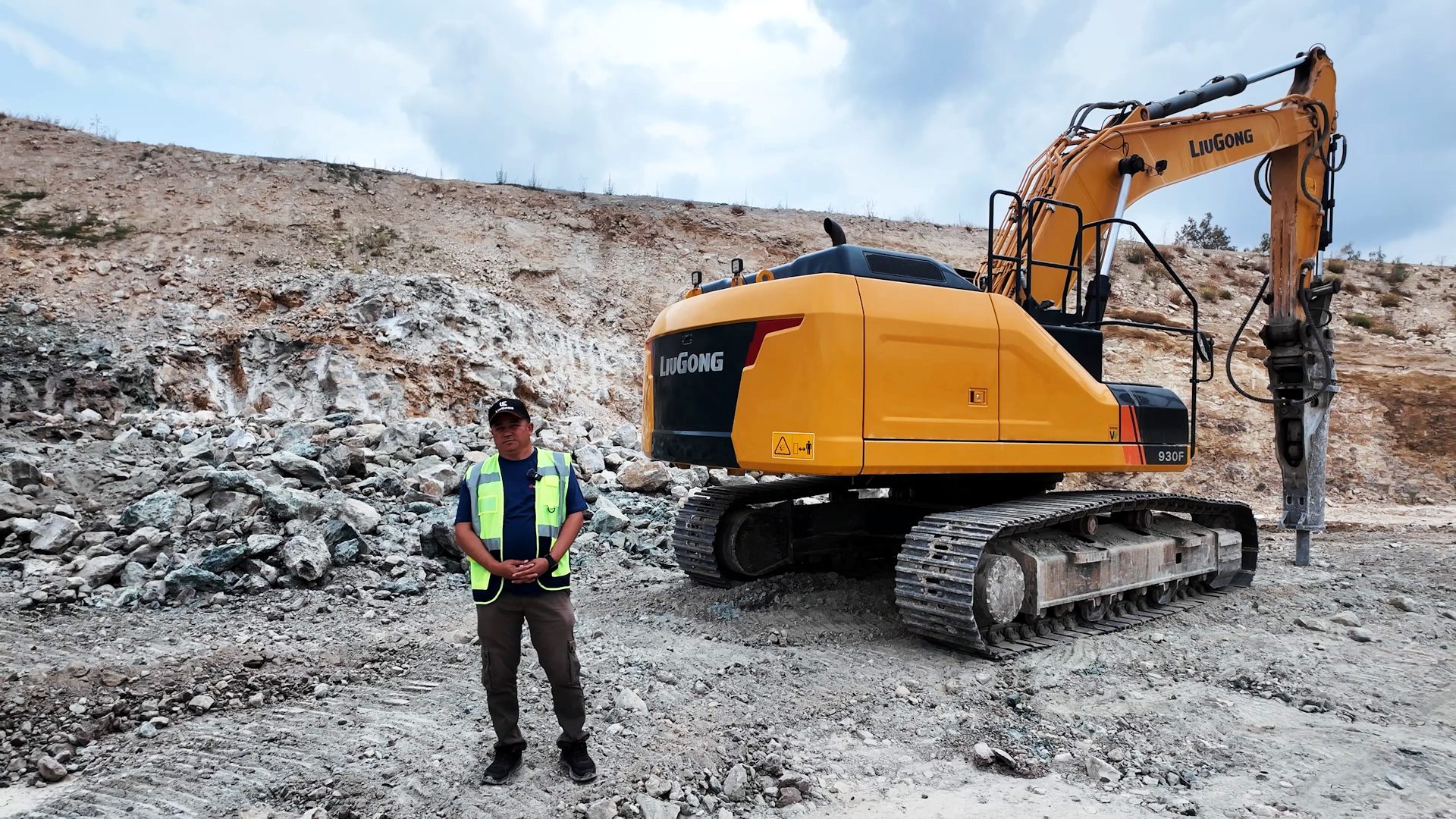 Man in safety vest with yellow excavator with breaker at a rocky site.