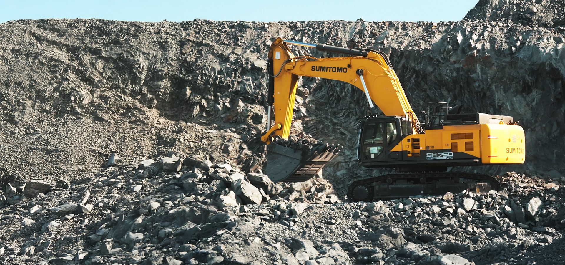 Yellow Sumitomo excavator digging in a rocky quarry.