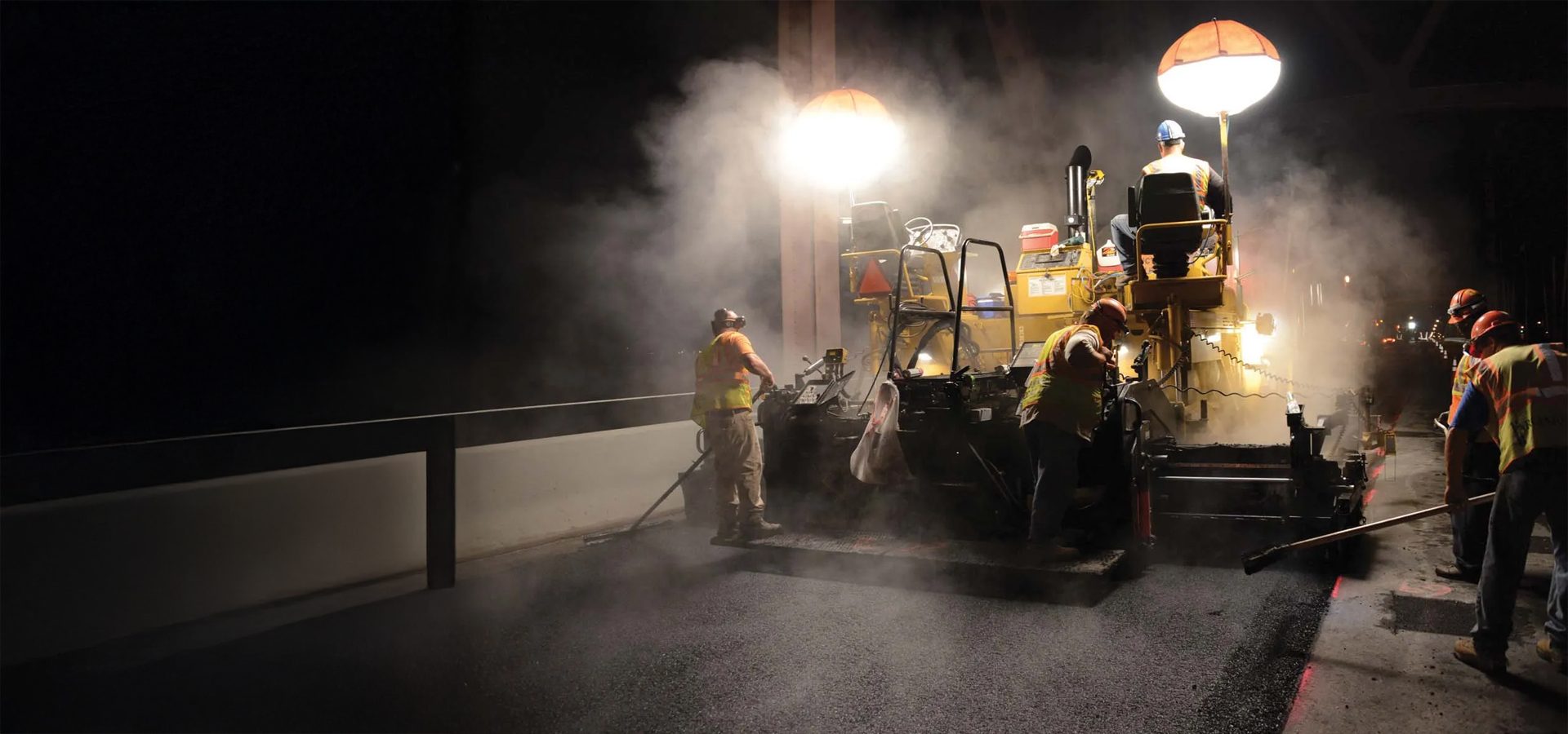 Workers use a large paving machine at night to lay asphalt, illuminated by bright lights, with steam rising.