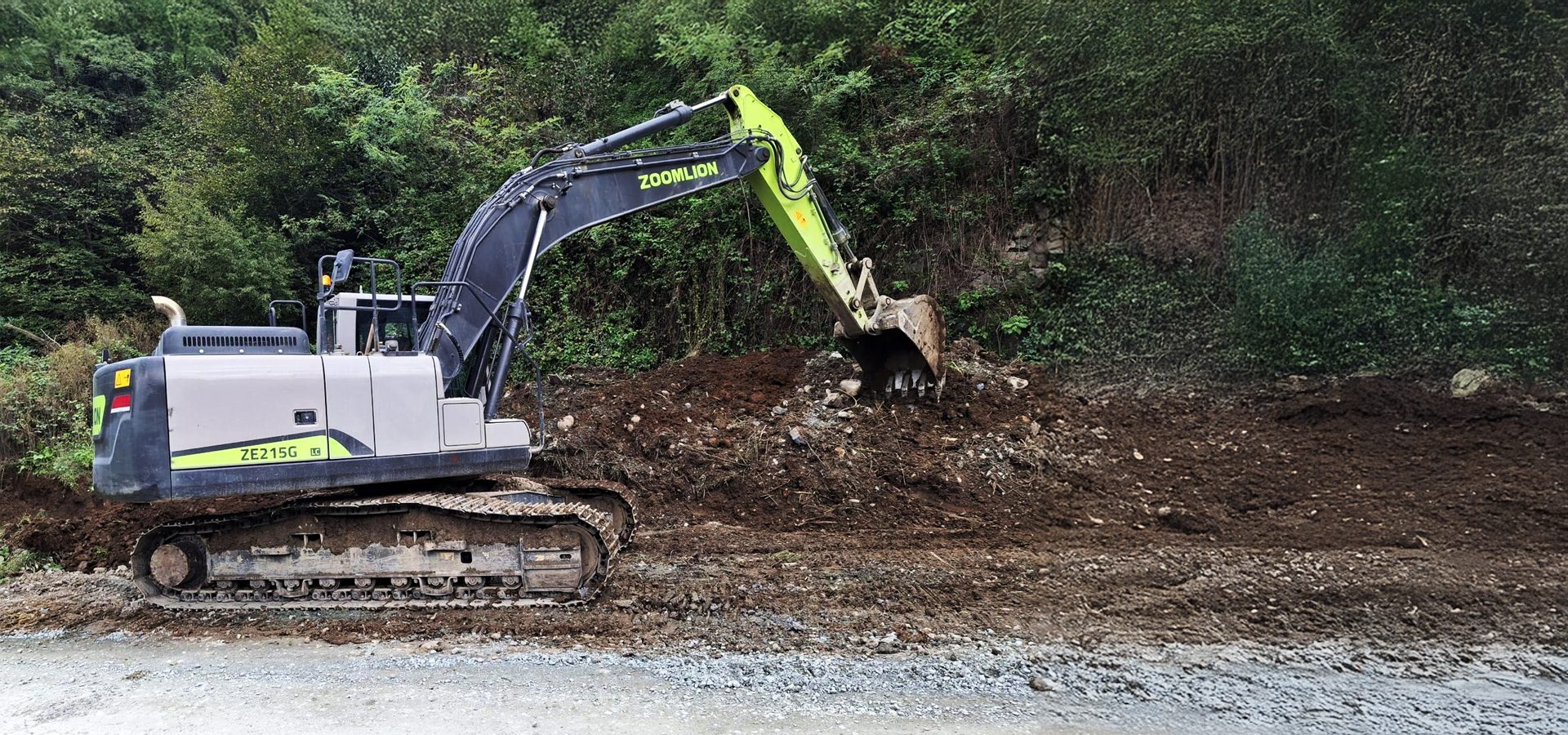 A ZOOMLION ZE215G excavator digging dirt next to a green, overgrown hillside.