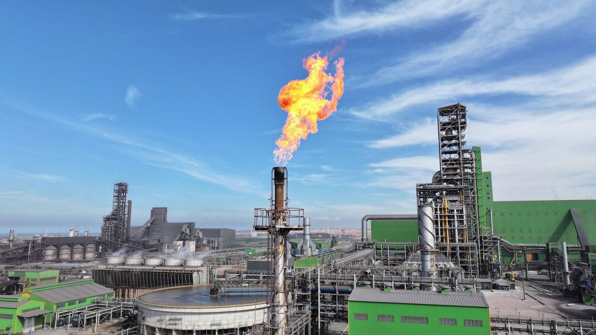 Aerial view of a large industrial complex with a prominent flare stack burning brightly under a blue sky.