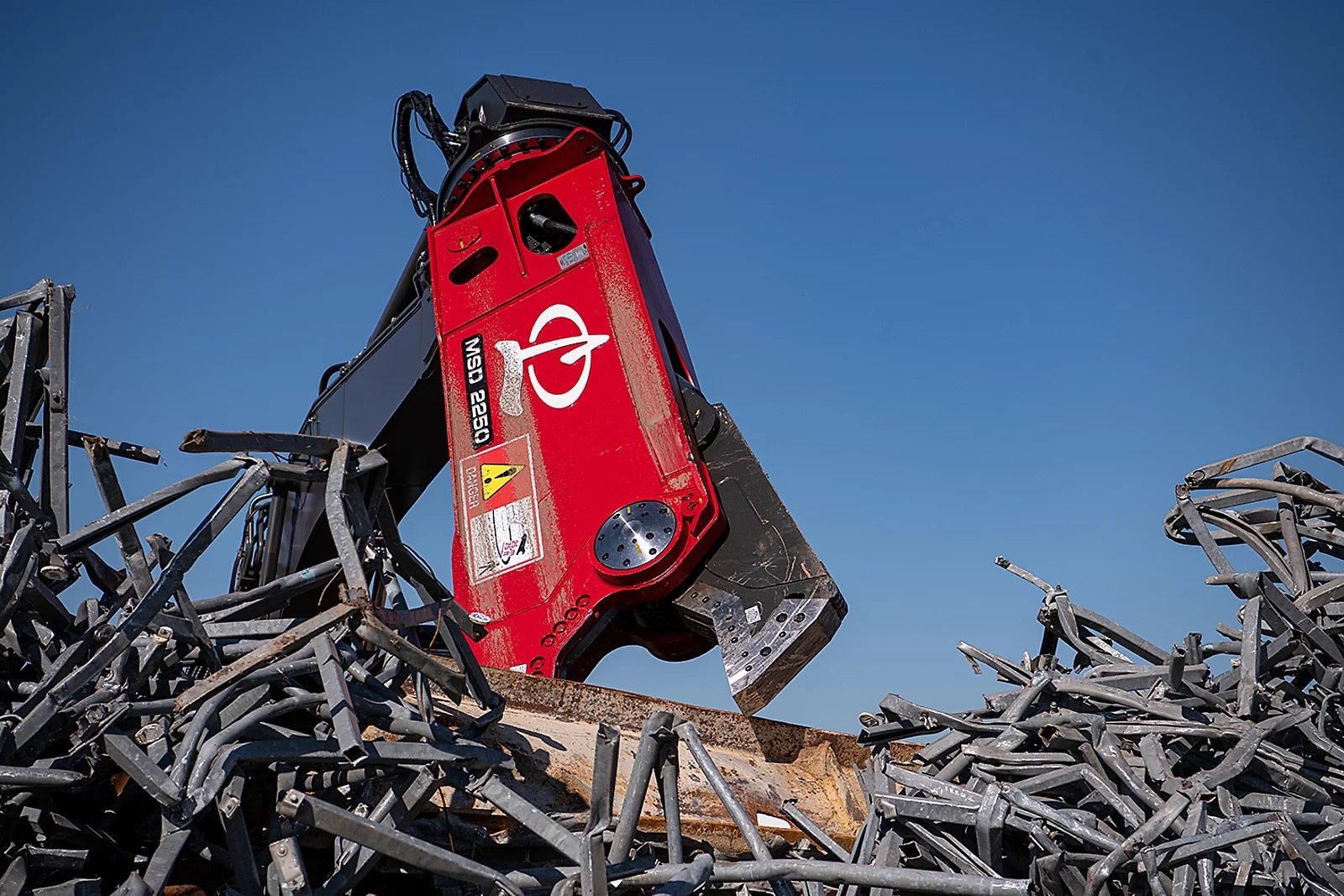 A large red hydraulic shear attachment processing a pile of grey scrap metal under a clear blue sky.