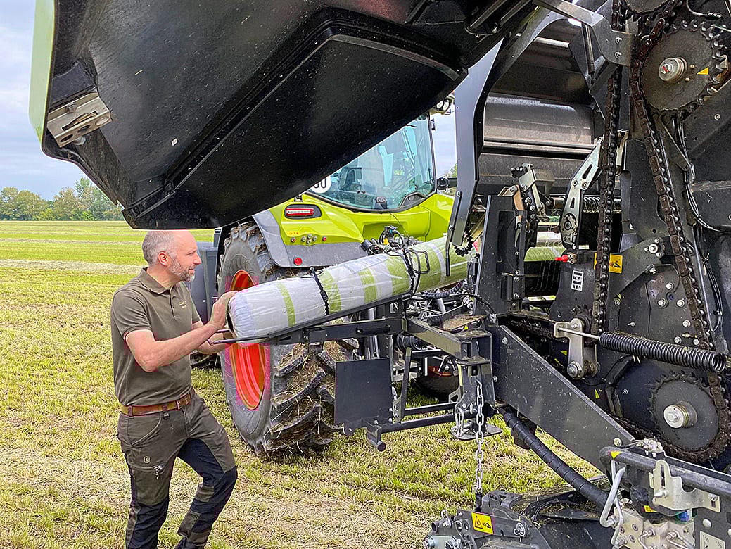 Man loading wrap into a baler machine in a field with a tractor in the background.