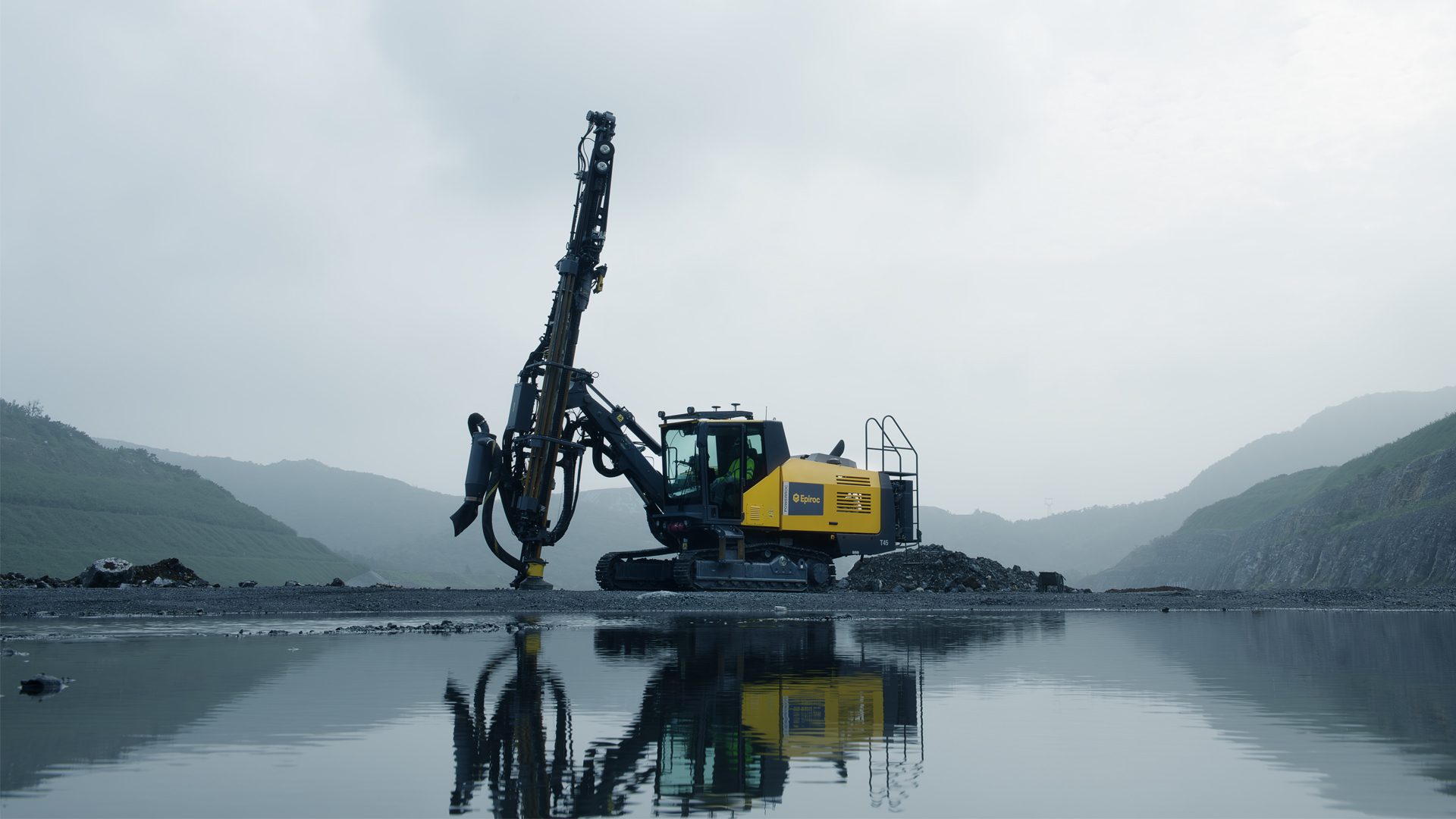 Large yellow drilling rig on a rocky shore, reflected in still water, with misty mountains.