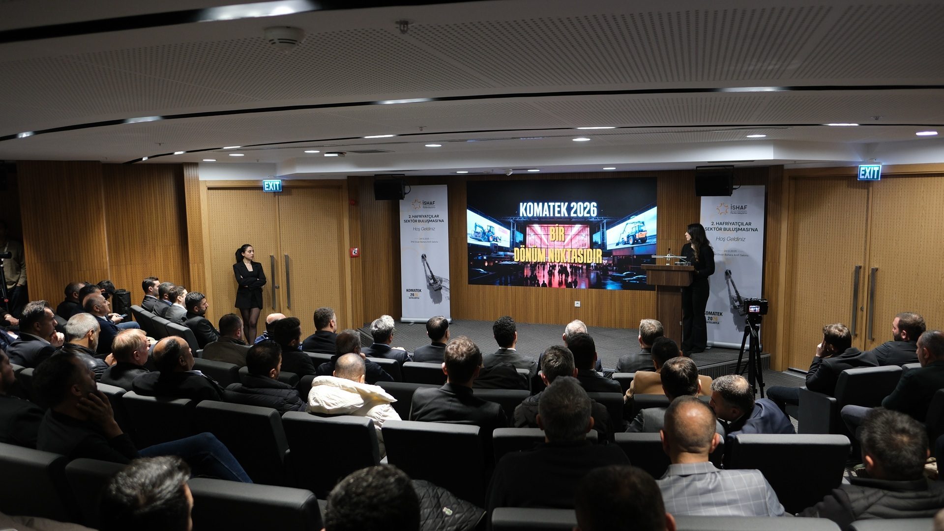 Conference with a speaker presenting KOMATEK 2026 to an audience in an auditorium.