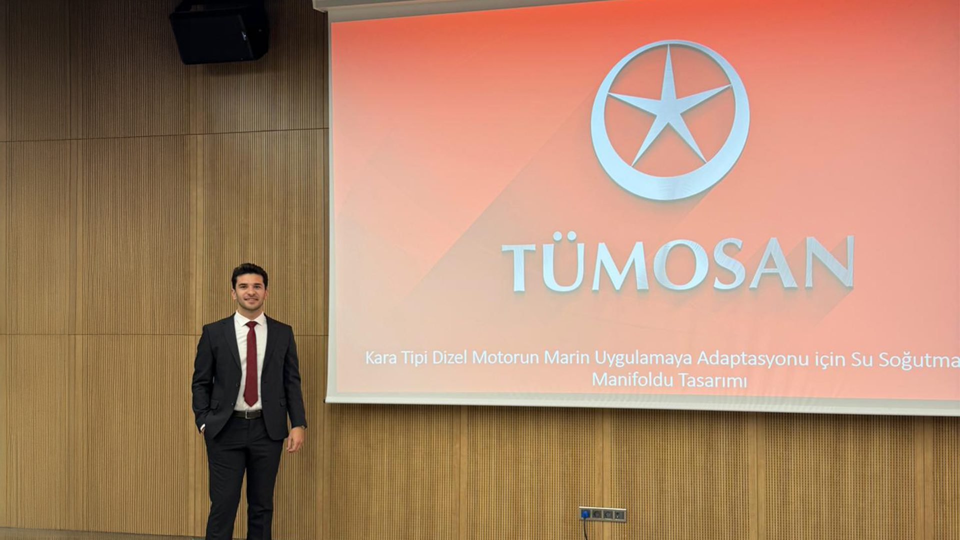A man in a suit stands by a screen displaying the "TÜMOSAN" logo and text about marine engine adaptation.