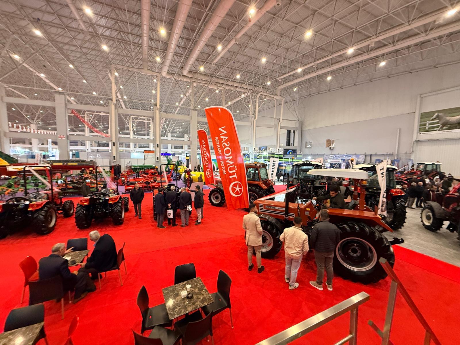 Indoor trade show featuring many tractors and agricultural equipment on a red floor, with people.