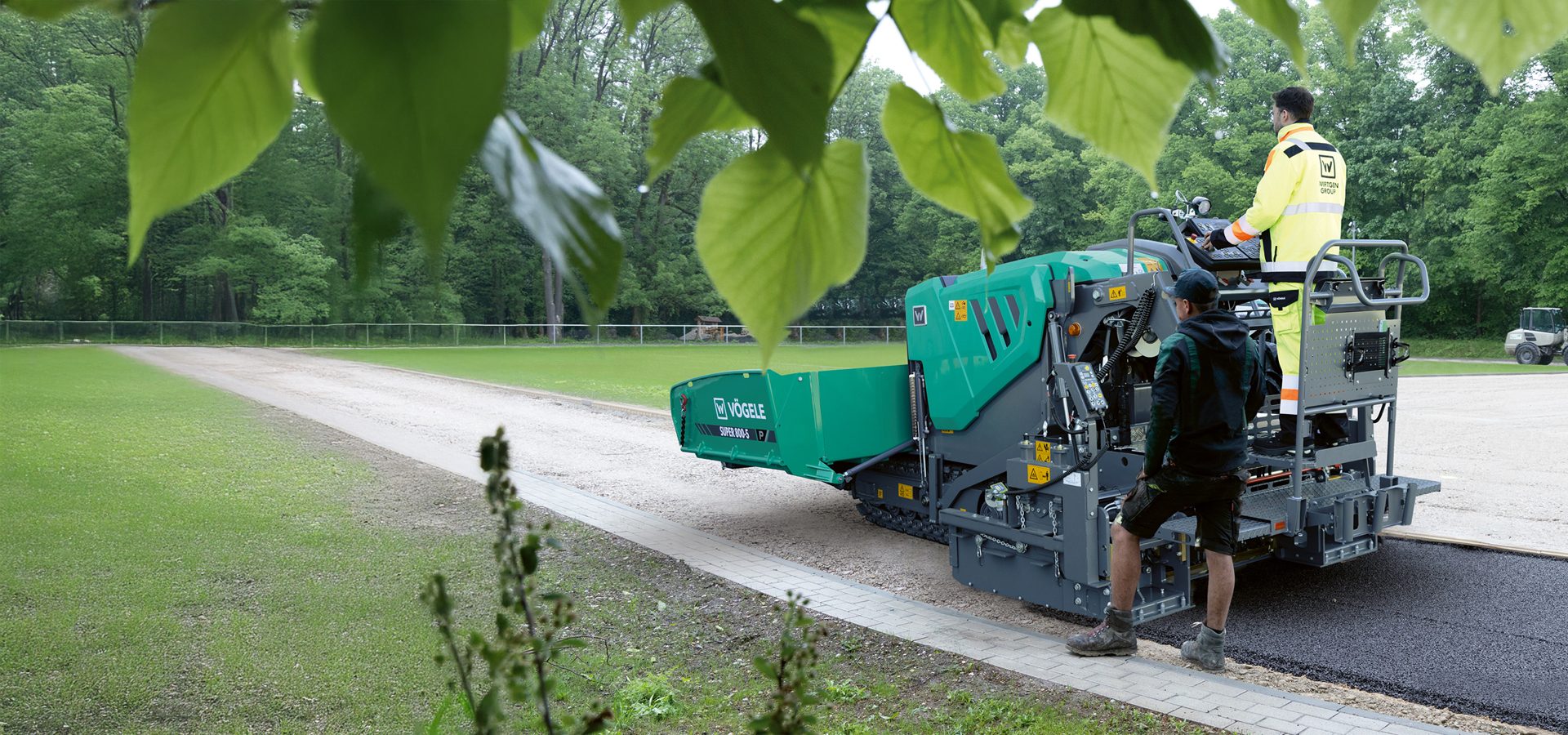 VÖGELE road paver lays asphalt with workers, framed by green leaves.