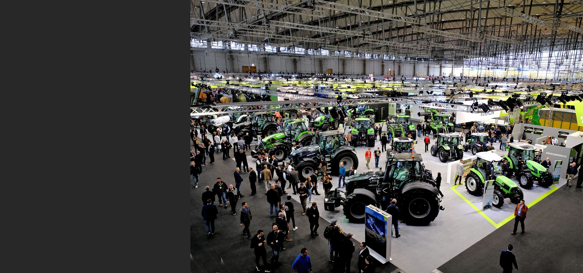 Indoor trade show with many green and black tractors on display and a crowd of people.