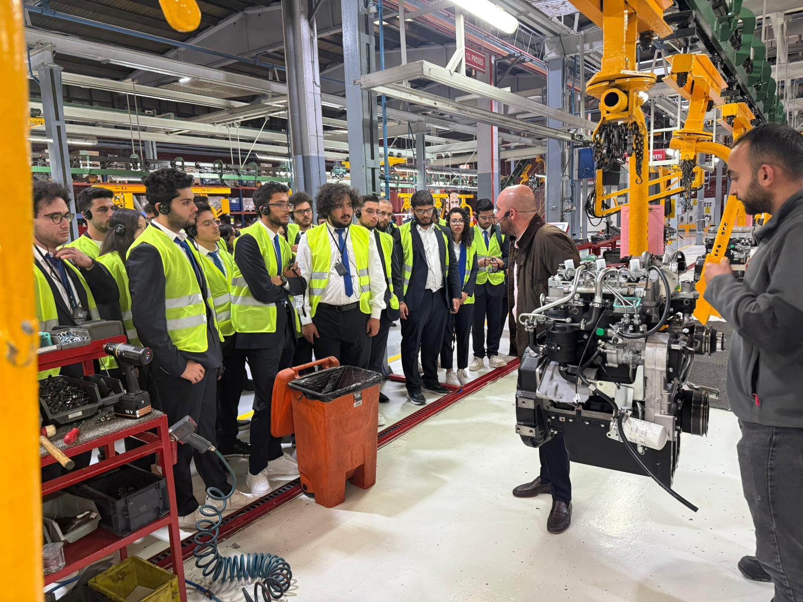 A group in safety vests listens to an instructor explaining an engine in a factory.