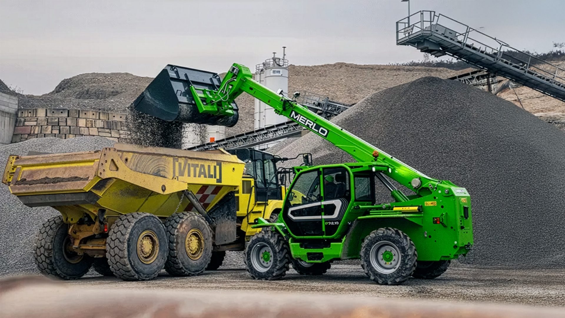 Green Merlo telehandler loads gravel into a yellow Vitali dump truck at a construction site.