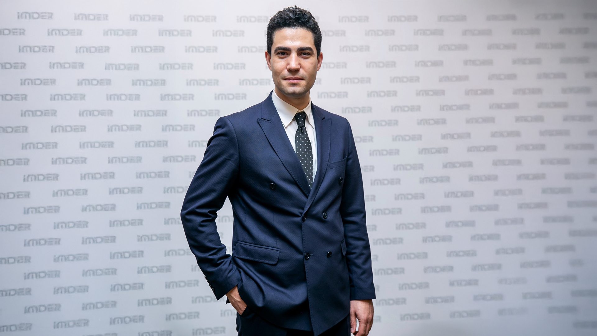 A man in a dark suit stands against a white wall patterned with repeating "IMDER" logos.