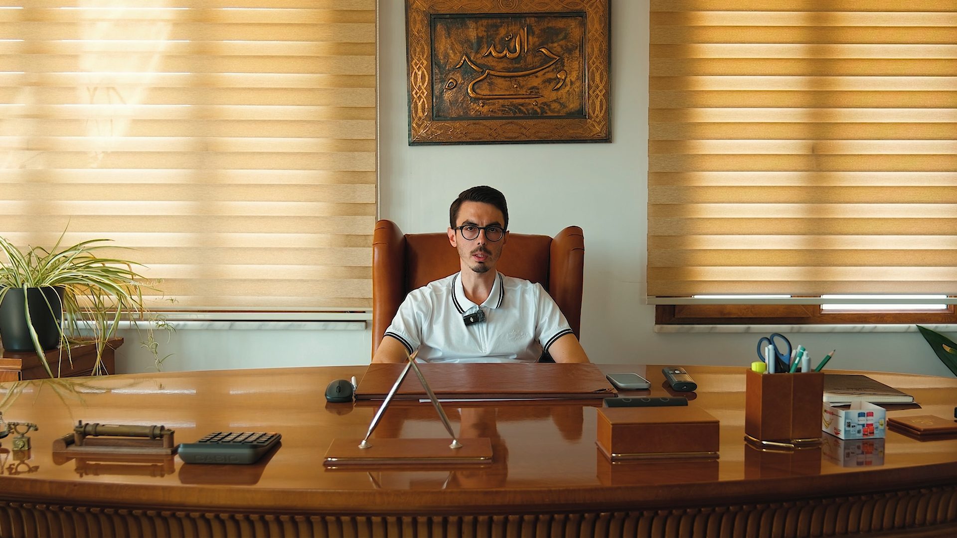 Man in glasses and white polo shirt sits behind a large wooden office desk with blinds and calligraphy art.