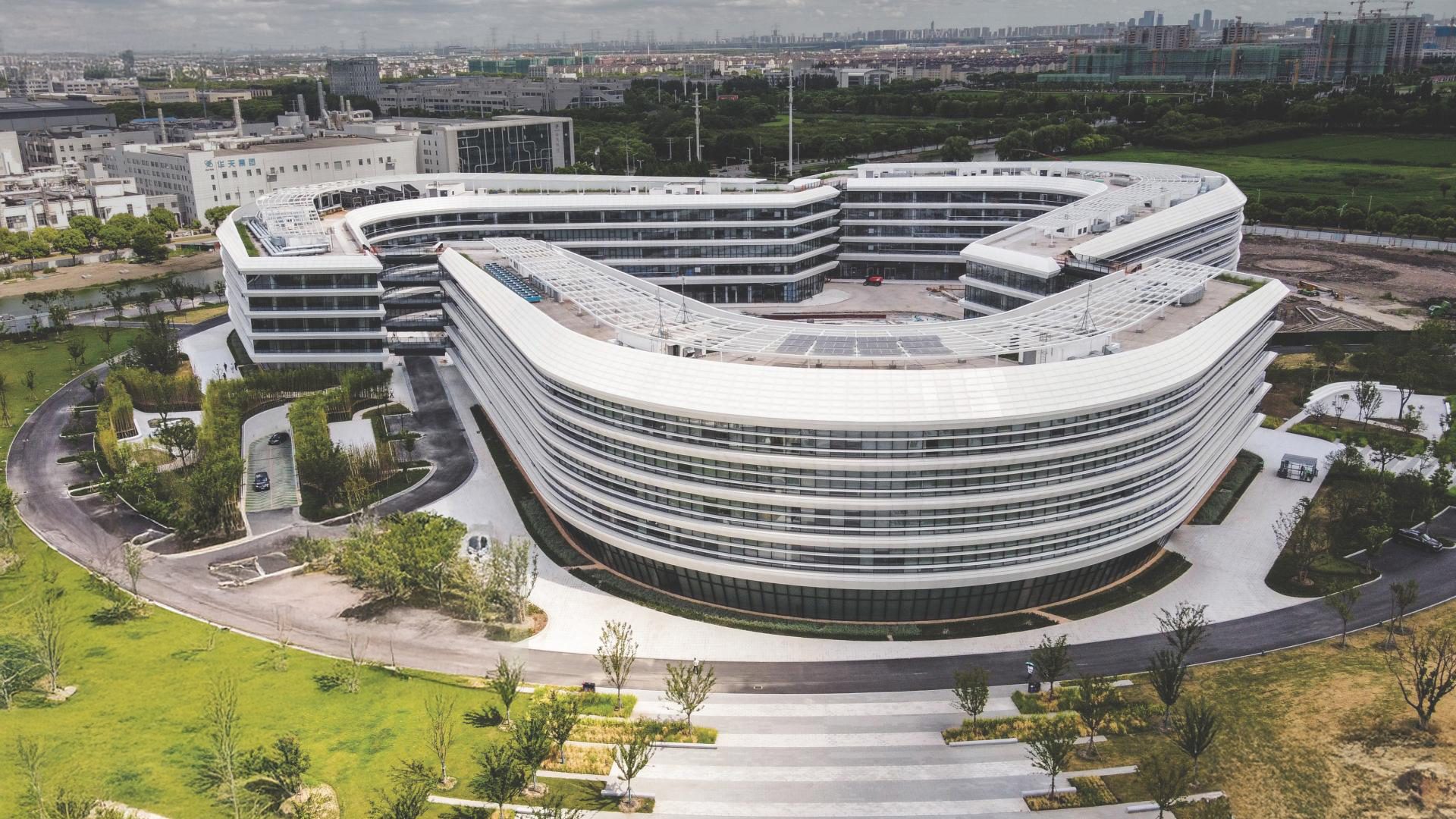 Aerial view of a modern, curved white building complex in a green landscape.