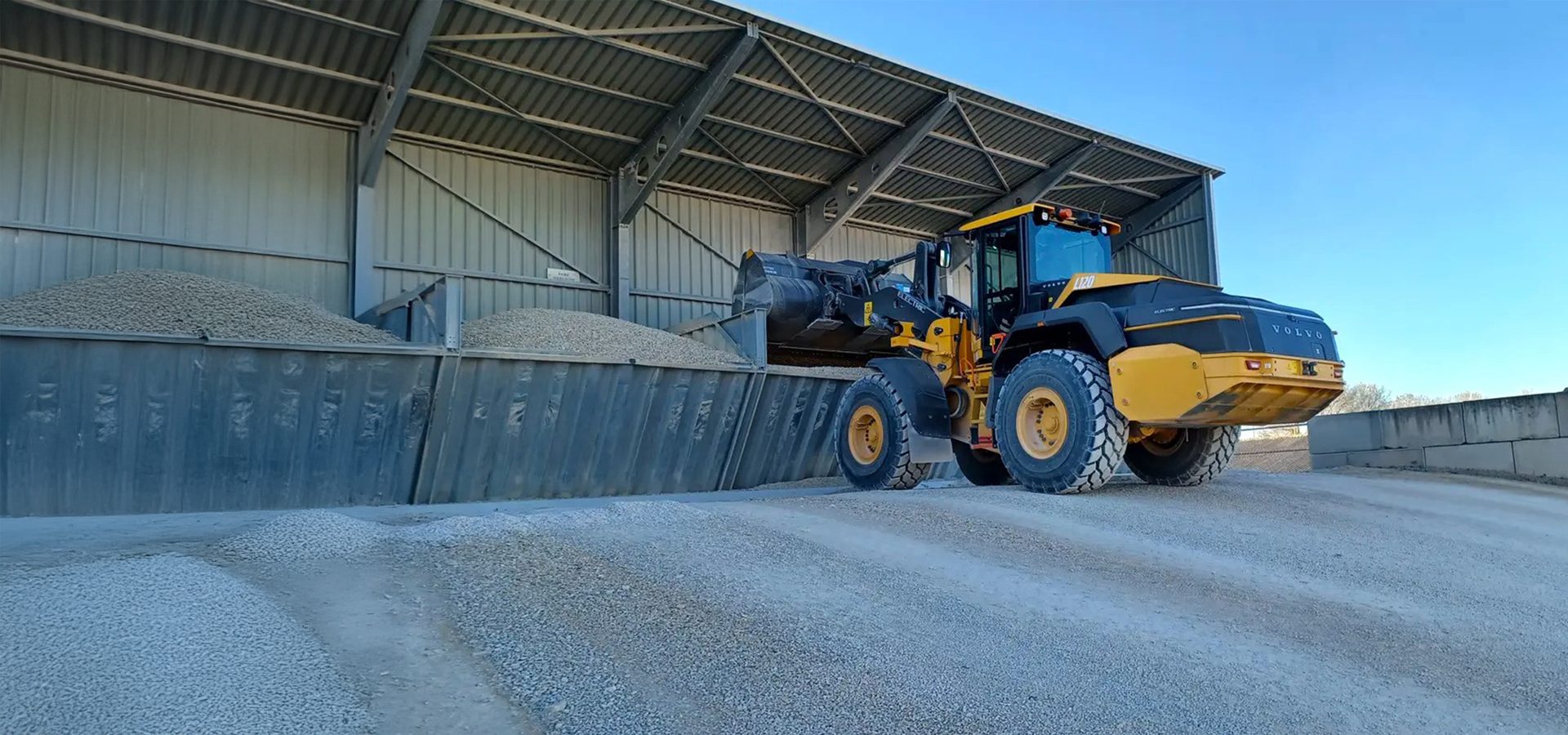 Yellow Volvo loader filling a bin with gravel under a shed.
