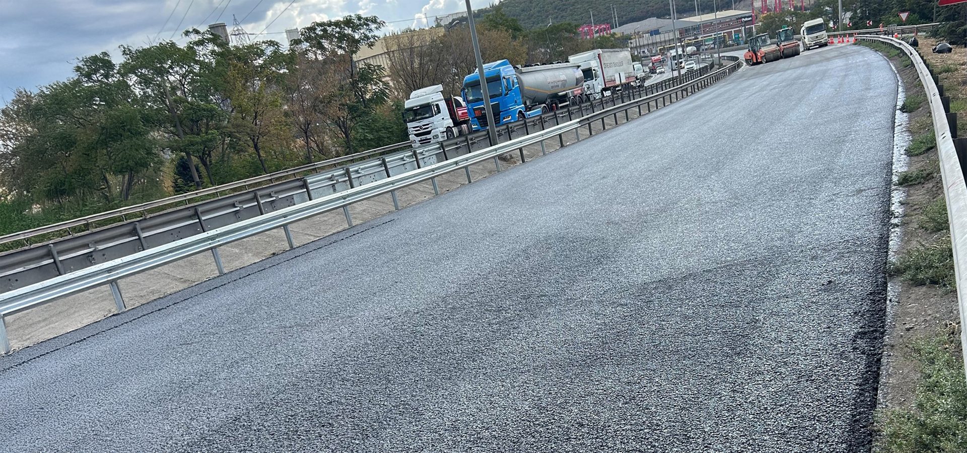 Newly paved asphalt road curving upwards, with heavy traffic and construction vehicles in the distance.