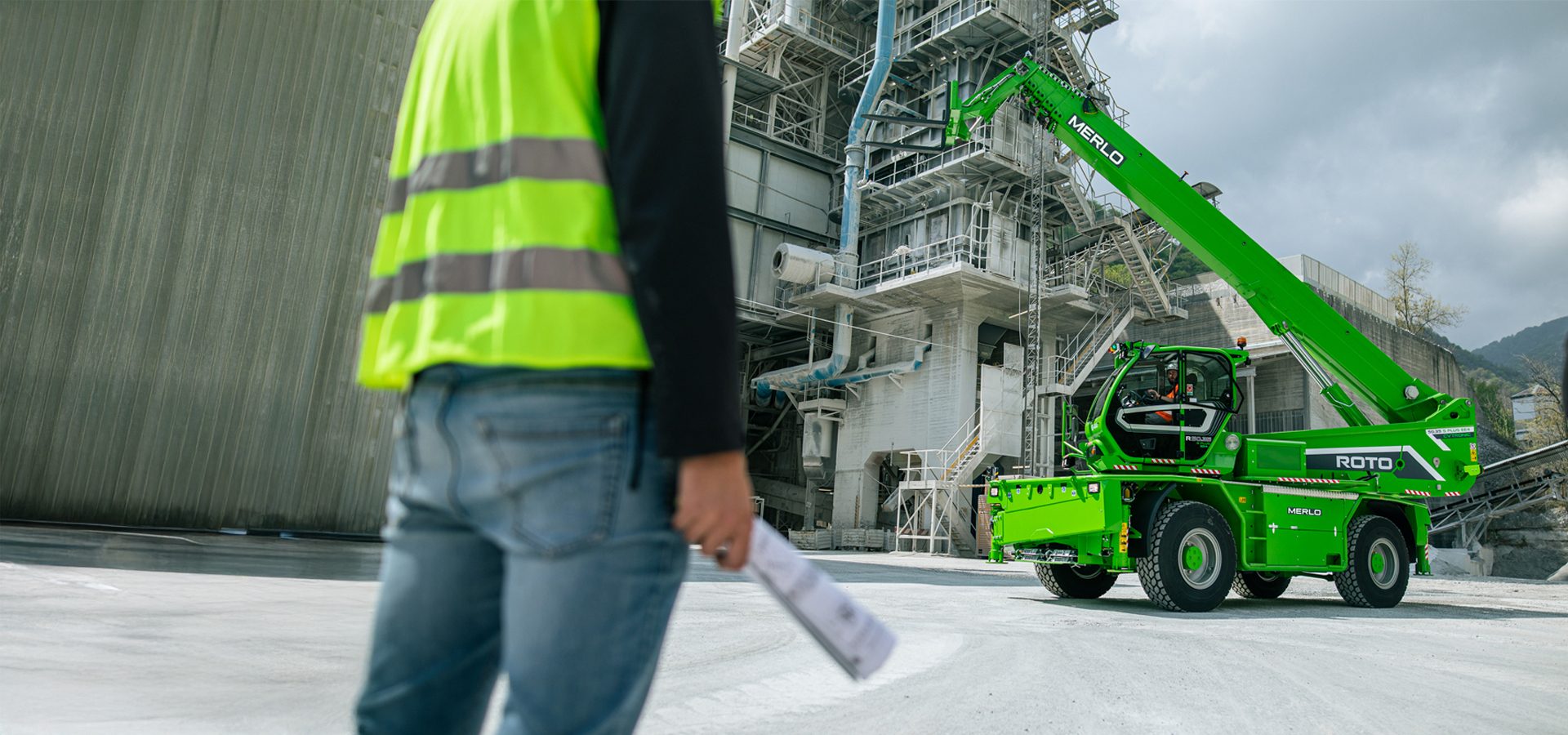 Worker in safety vest looks at green Merlo telehandler at an industrial plant.