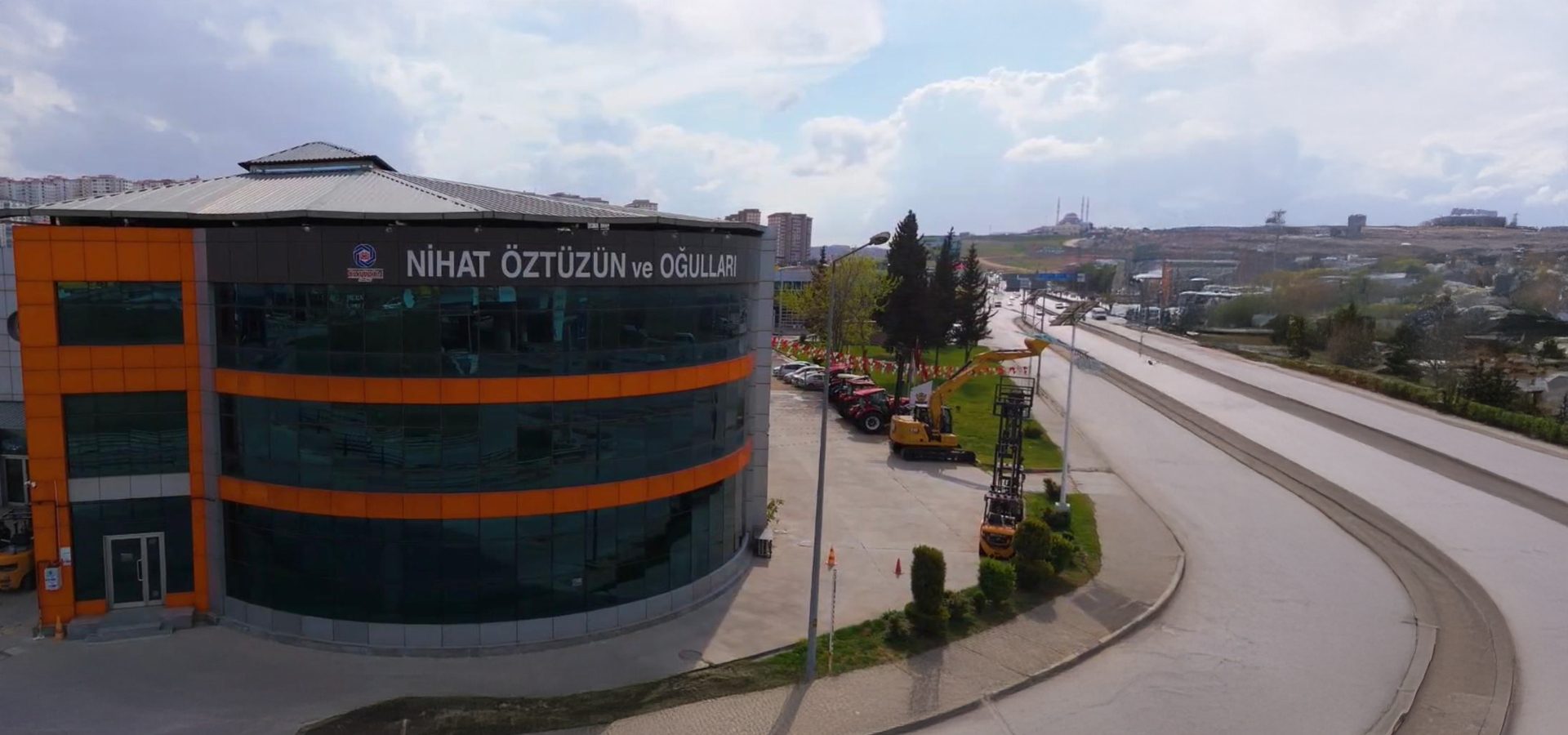 Modern building with "Nihat Öztüzün ve Oğulları" sign, heavy machinery outside, and a distant mosque.