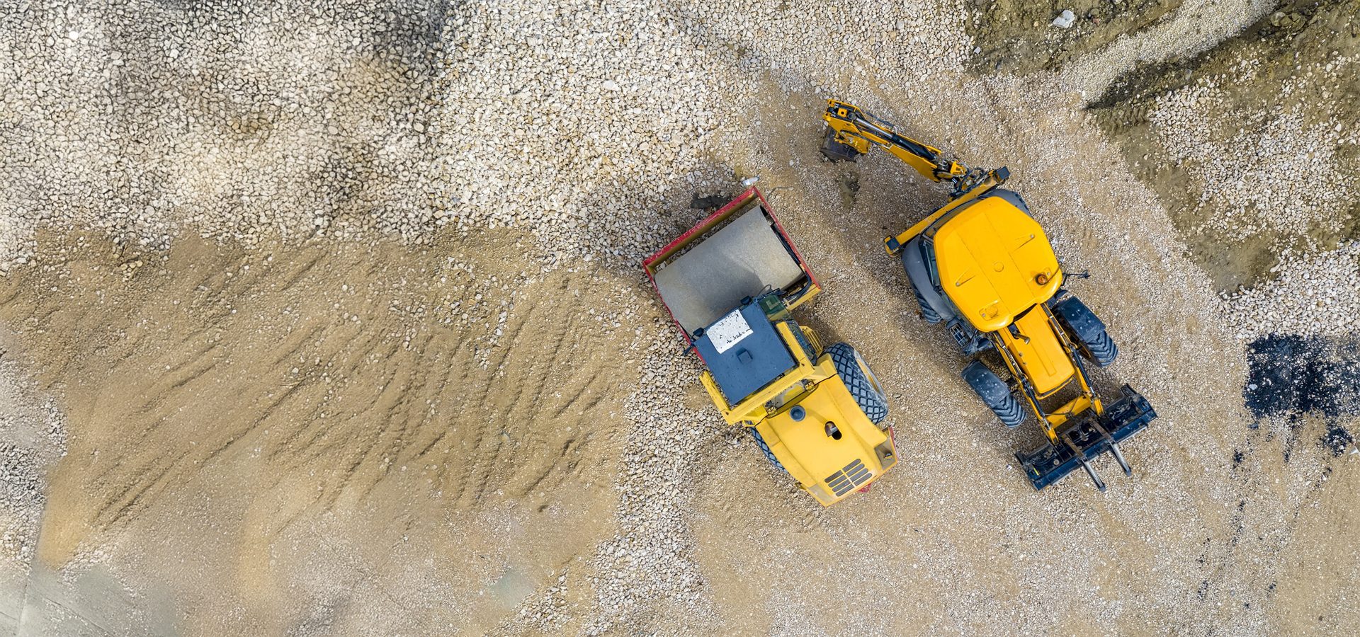 Aerial view of a construction site with two yellow heavy machinery vehicles, a dump truck and an excavator, on gravel and dirt.